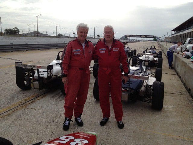 Two men in red suits standing next to a race car.