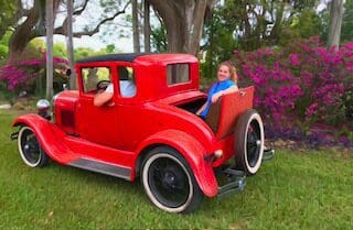 A woman sits in a red Ford Model A car at Daytona Speedway.